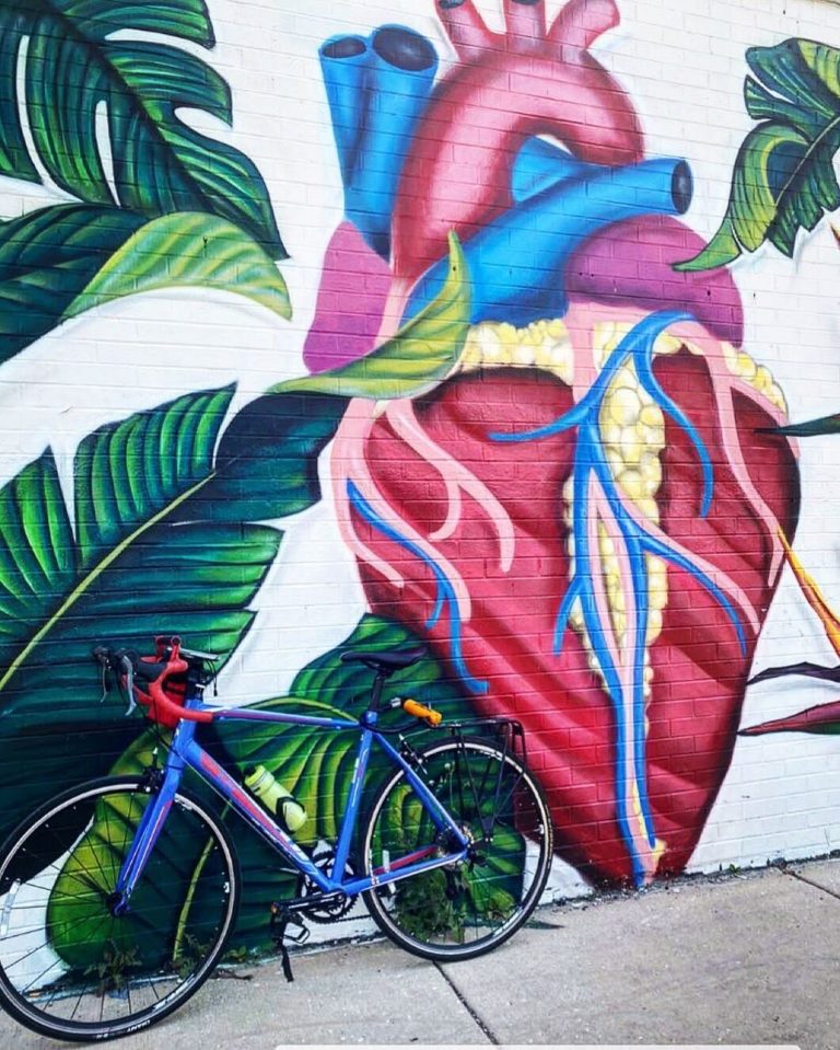 A blue bike with drop bars lying against a wall mural of a heart.